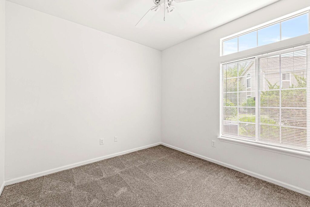 Empty room with white walls, carpeted floor, ceiling fan, and large window letting in natural light.