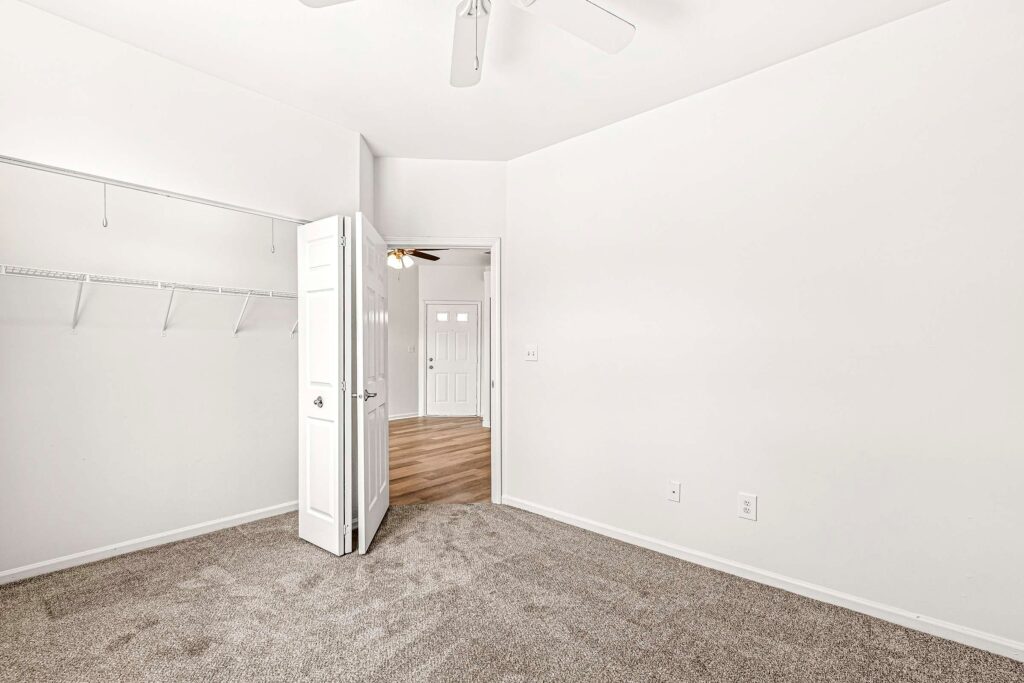 Empty room with white walls, beige carpet, an open closet with wire shelves, and an open door leading to a hallway with wood flooring and a white entrance door.