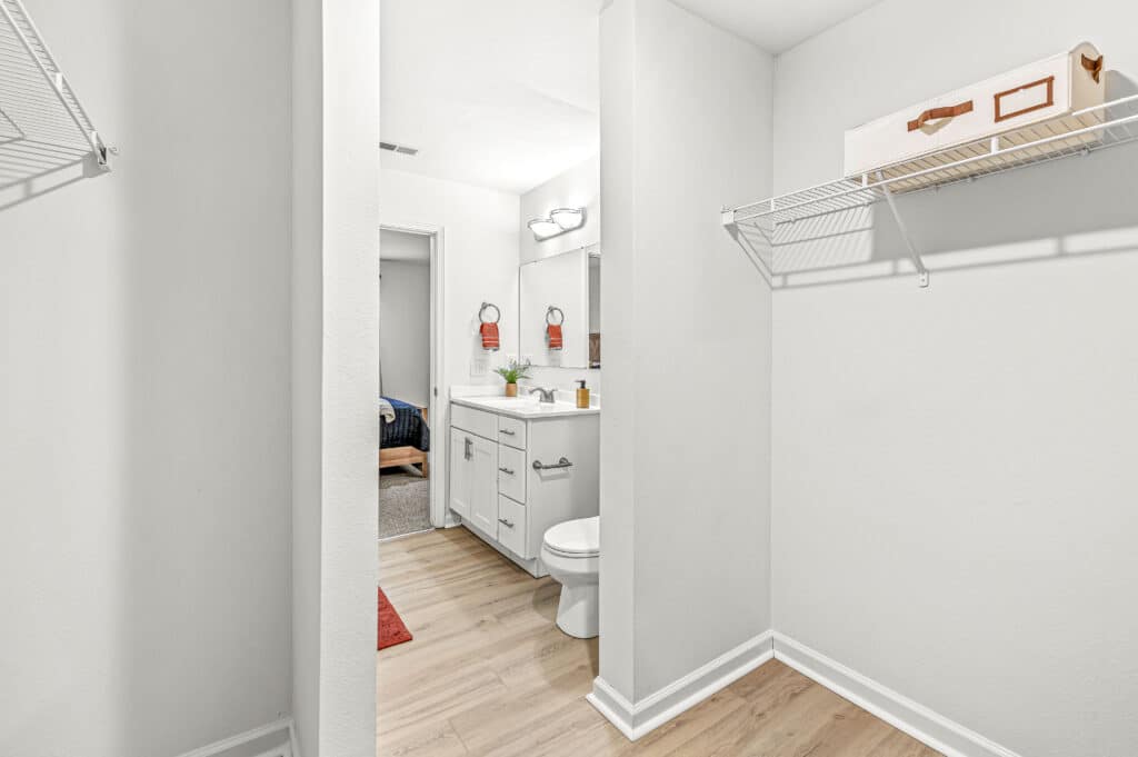 Enjoy a bright white bathroom at Arborwood Apartments, featuring a vanity, mirror, toilet, and open shelving beside a closet with wire shelves and a box. Light wood flooring flows seamlessly throughout the space.