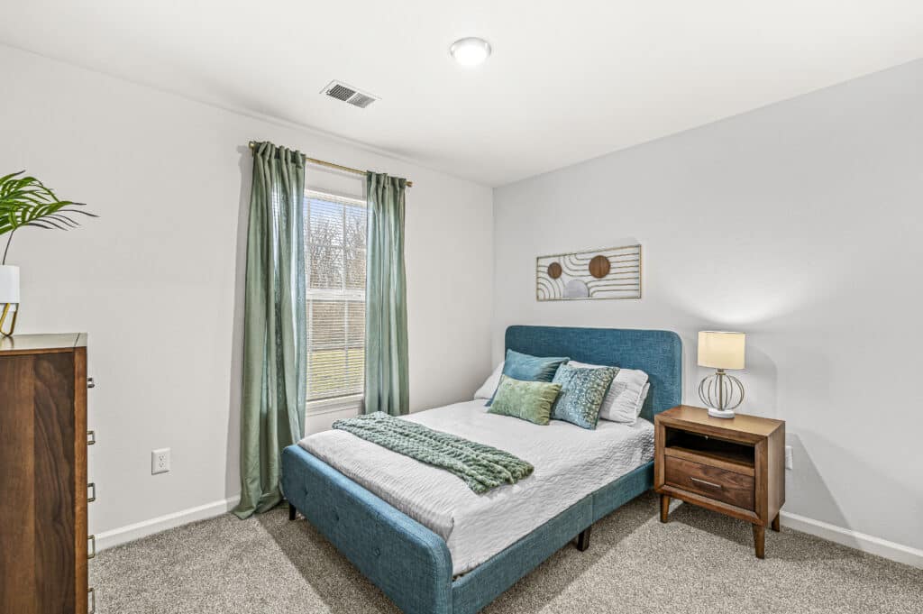 A bedroom at Arborwood Apartments features a teal upholstered bed, white bedding, two green pillows, a wooden nightstand with a lamp, a dresser, and a window adorned with green curtains.