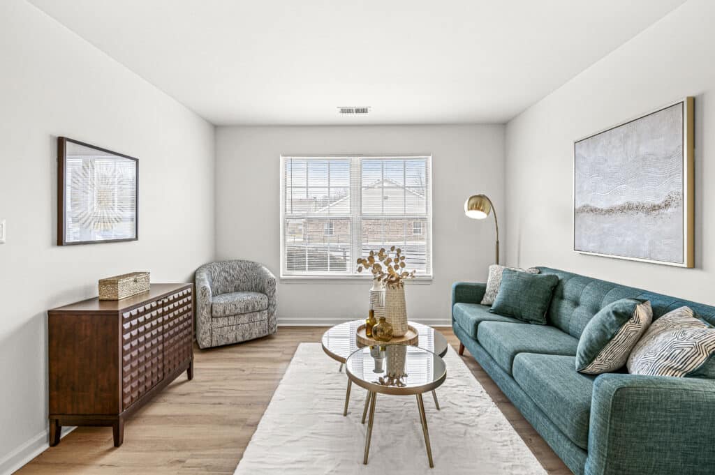 Bright living room at Arborwood Apartments with a teal sofa, round glass coffee tables, an armchair, wooden cabinet, and a large window letting in natural light.