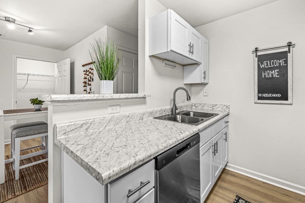 Modern kitchen at Arborwood Apartments featuring white cabinets, marble countertop, stainless steel sink, dishwasher, and a decorative plant; a chalkboard sign on the wall reads "Welcome Home.