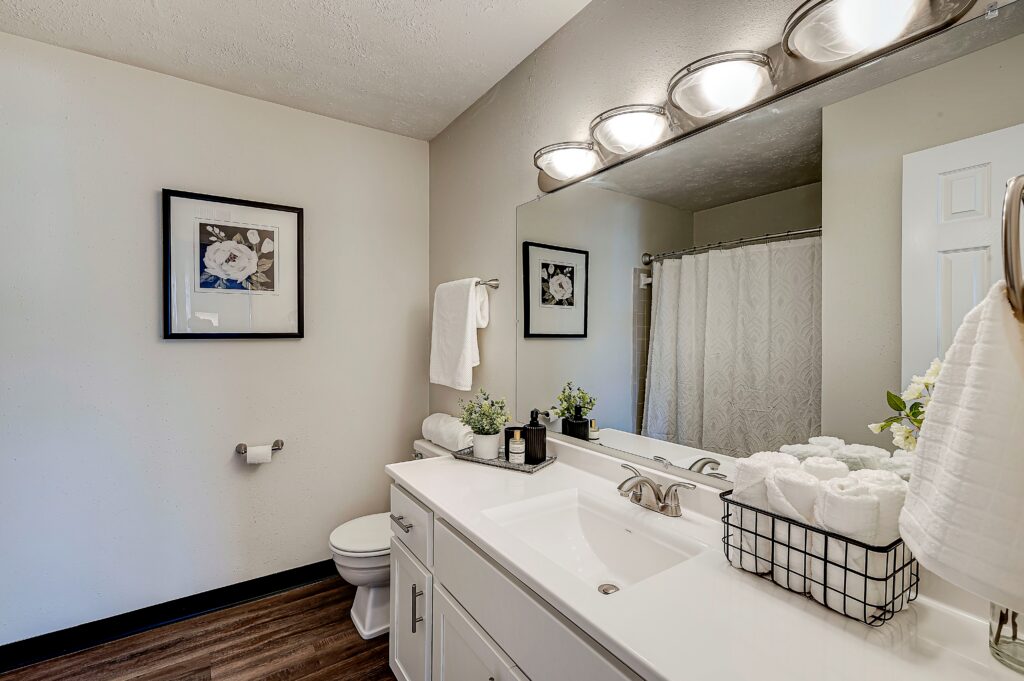 Modern bathroom at Estates of Creekside Apartments featuring a white vanity, large mirror, double sink, rolled towels in a wire basket, stylish wall art, and a shower with a white curtain.