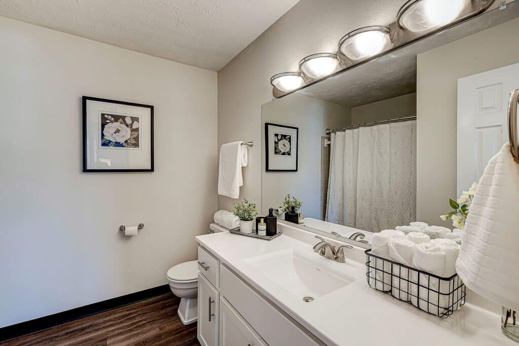 Modern bathroom with white vanity, large mirror, basket of rolled towels, wall art, toilet, and shower with a white curtain; wood-look flooring and neutral walls.
