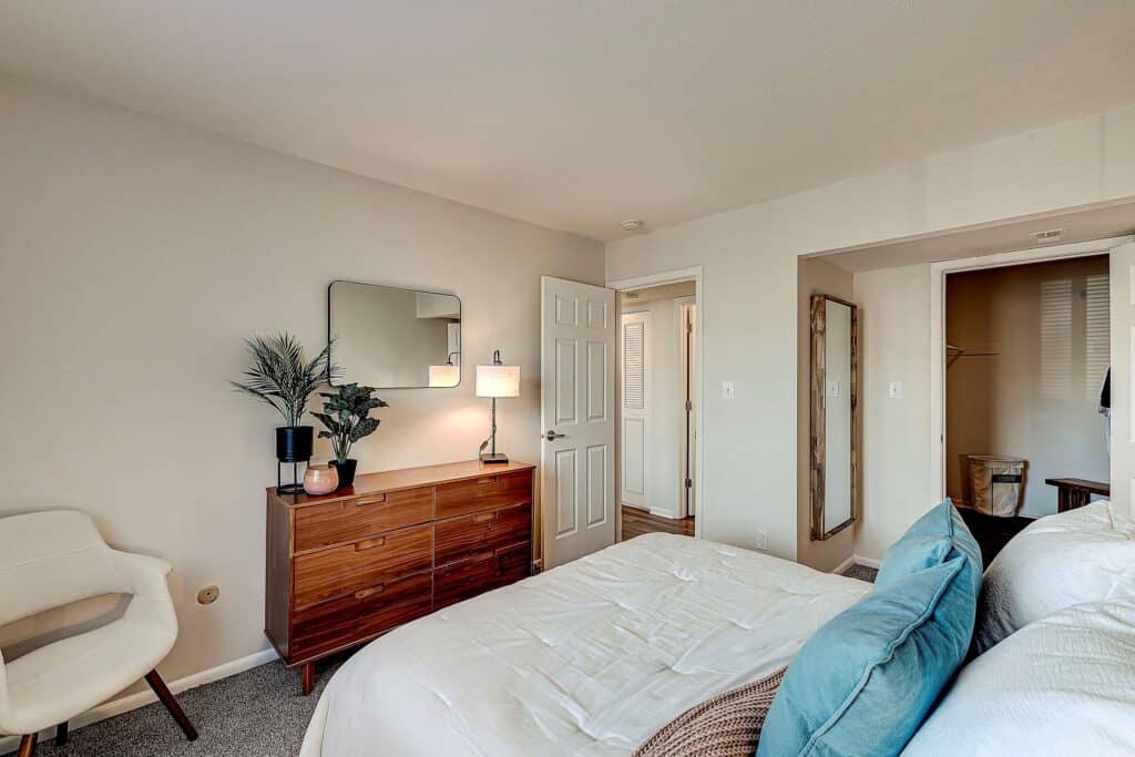 A neatly arranged bedroom with a bed, a wooden dresser with a mirror, a white chair, a standing lamp, and an open door leading to a hallway and closet.