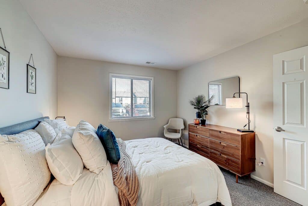 A neatly made bed with white bedding and blue pillows is in a small bedroom with a window, wooden dresser, chair, and neutral decor.