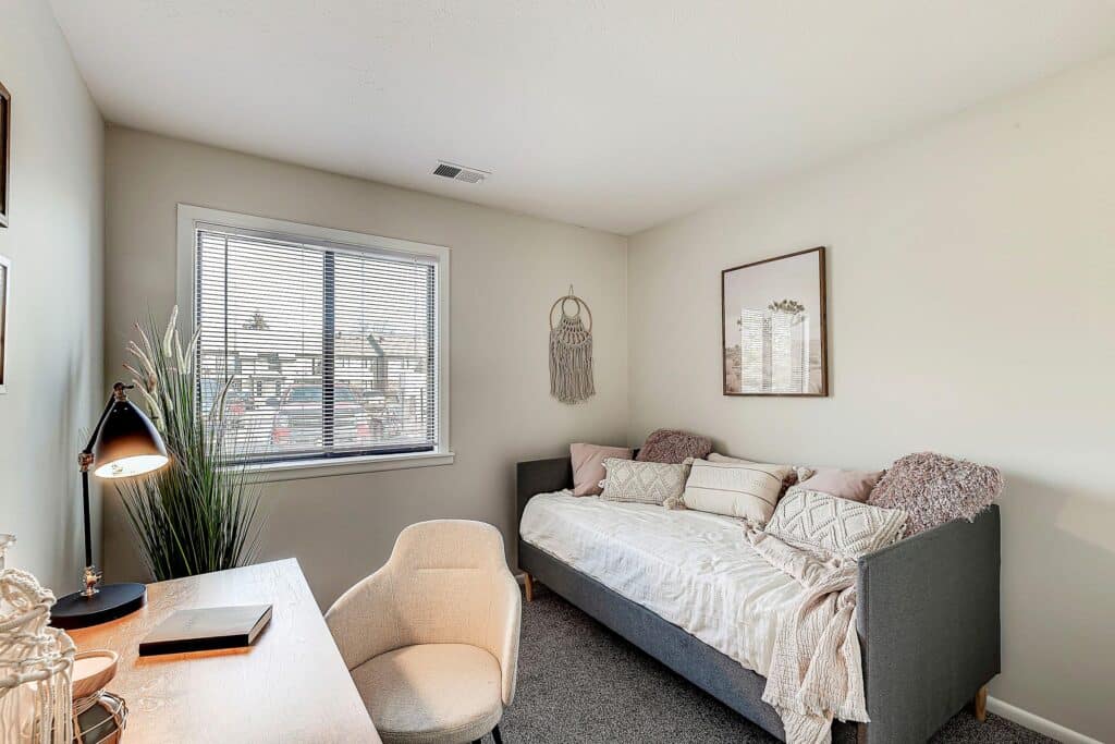 A small home office with a desk and chair, a daybed with pillows and throw blanket, a window with blinds, and minimal wall decor.