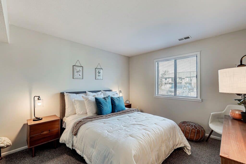 A neatly made bed with blue accent pillows is centered in a bedroom with neutral walls, two nightstands, a window with blinds, and minimal decor.