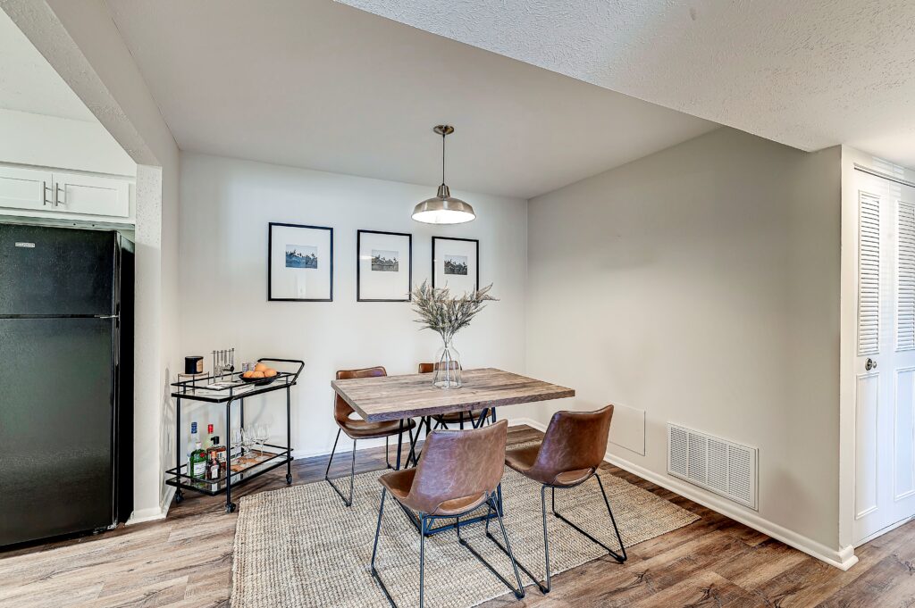 A modern dining area at Estates of Creekside Apartments features a wooden table, four brown chairs, a light fixture overhead, a bar cart, and framed pictures on the wall.