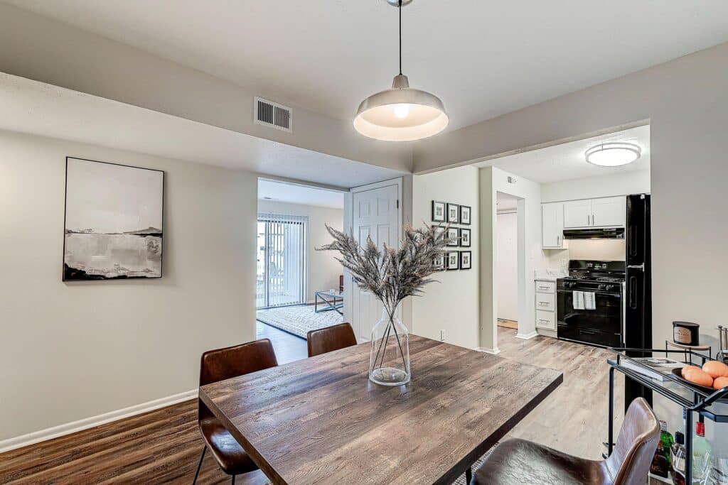 A modern dining area with a wooden table, four brown chairs, a vase with dried flowers, and a hanging light fixture, adjacent to a kitchen and living room.