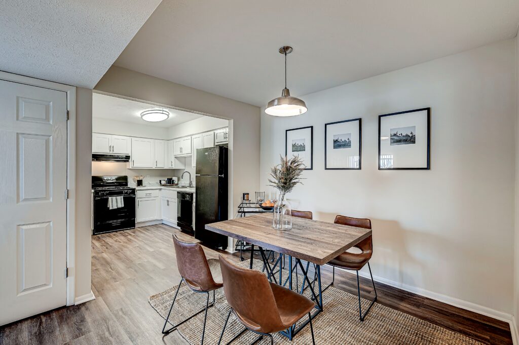 A modern dining area at Estates of Creekside Apartments features a wooden table, four brown chairs, and a vase with dried plants, adjacent to a stylish kitchen with white cabinets and black appliances.
