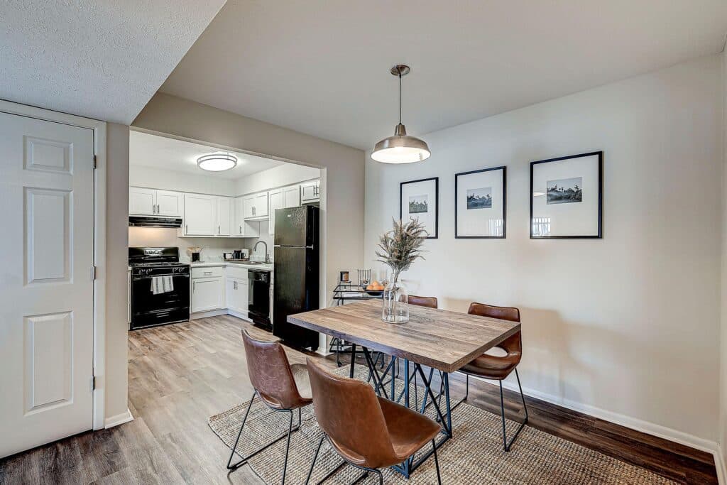 A dining area with a wooden table and four brown chairs is adjacent to a kitchen with white cabinets and black appliances. Three framed photos hang on the wall above the table.