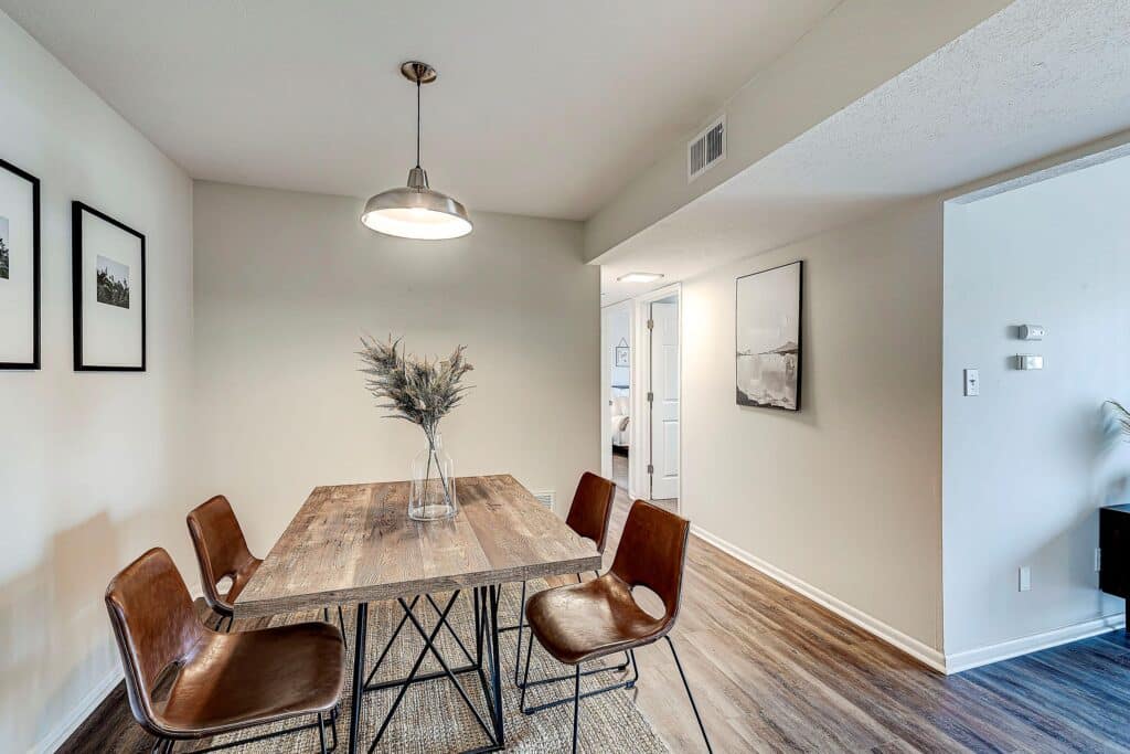 A dining area with a wooden table, four brown chairs, a vase with dried branches, wall art, and a ceiling light in a modern, neutral-toned room.
