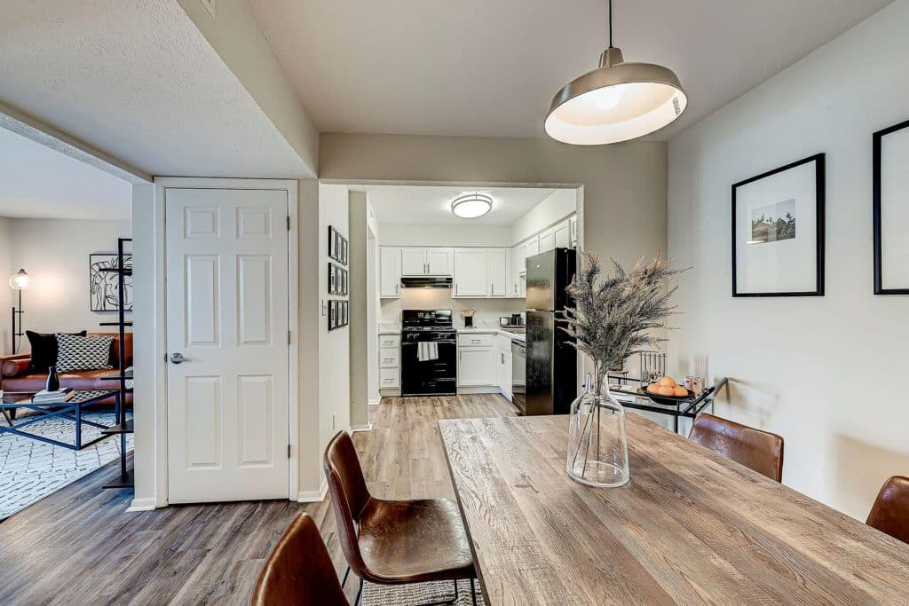 A dining area with a wooden table and brown chairs sits next to a kitchen with white cabinets, black appliances, and an adjacent living room with a sofa and decor.