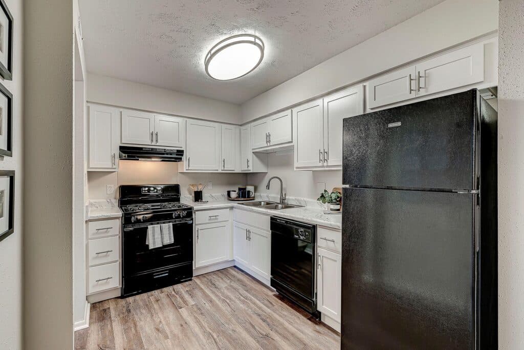Modern kitchen with white cabinets, black appliances including stove, dishwasher, and refrigerator, marble countertops, and wood-style flooring under a round ceiling light.
