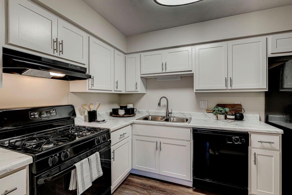 Modern kitchen with white cabinets, black appliances, gas stove, double sink, and marble countertops. Cooking utensils and small appliances are on the countertop.