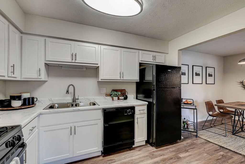 Modern kitchen with white cabinets, black appliances, double sink, and a dining area with a table and four chairs in the background.