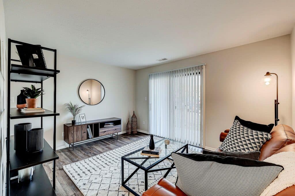 Modern living room with a leather sofa, geometric rug, black shelving unit, TV stand, round mirror, and sliding glass doors letting in natural light.