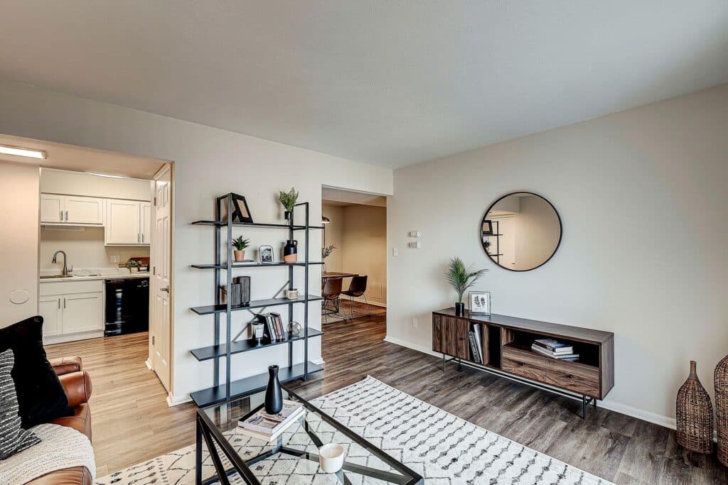 Modern living room with wood flooring, a glass coffee table, open shelving, media console, round mirror, and a view into a kitchen and dining area. Neutral decor and natural light.