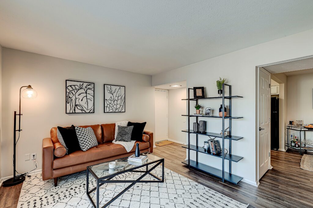 A modern living room at Estates of Creekside Apartments features a brown sofa, black-and-white decor, a glass coffee table, a bookshelf, and a patterned rug atop stylish wood flooring.