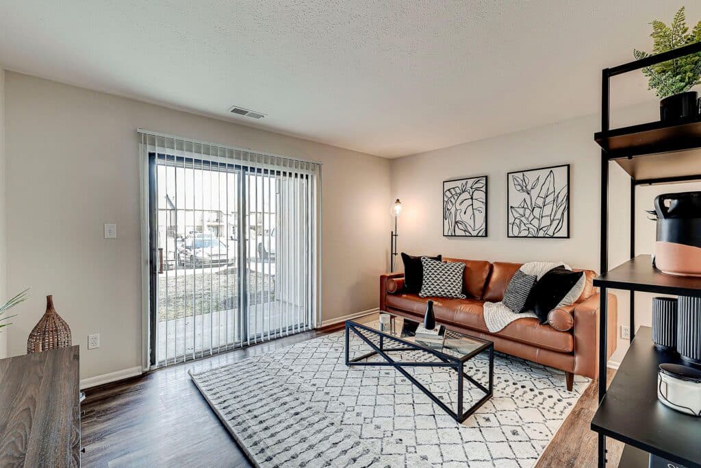 Modern living room with a brown leather sofa, glass coffee table, patterned rug, wall art, and sliding glass doors leading outside. Natural light brightens the space.