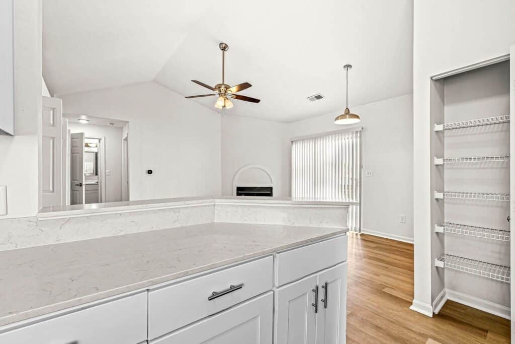 Modern kitchen with white cabinets and marble countertops, open to a living area with ceiling fan, pendant light, built-in shelves, and sliding glass doors with vertical blinds.