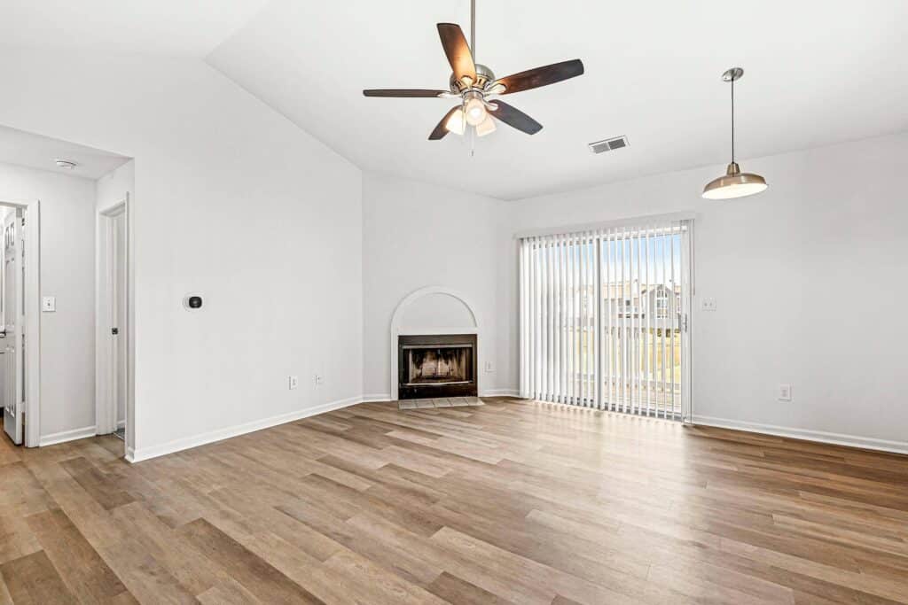 Unfurnished living room with wood flooring, ceiling fan, fireplace, pendant light, and sliding glass doors leading to a patio. Walls are white and space is well-lit by natural light.