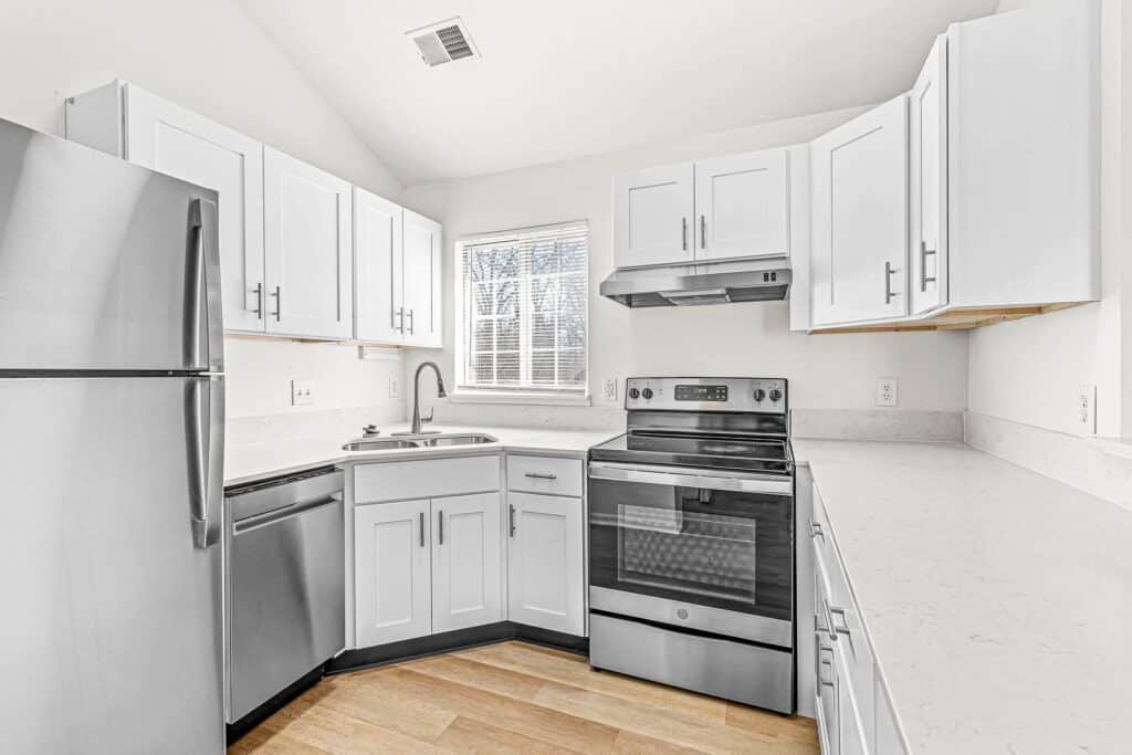 Modern kitchen with white cabinets, stainless steel appliances, electric stove, dishwasher, refrigerator, and a window above the sink. Light wood flooring and quartz countertops.