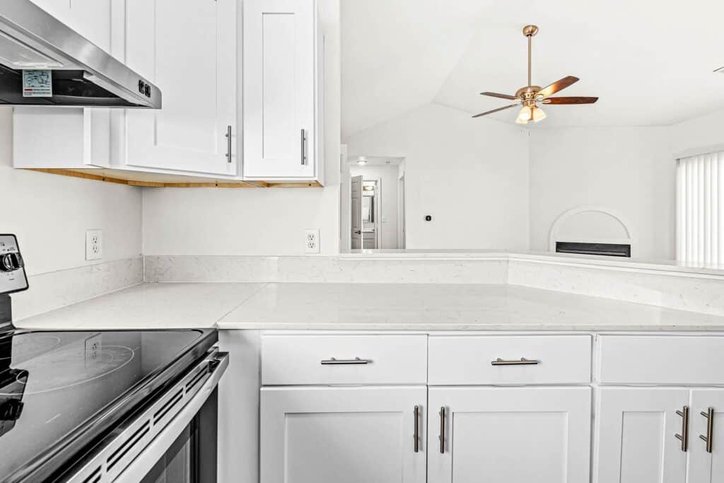 Modern kitchen with white cabinets, stainless steel stove, marble countertops, and a view into a bright living area with a ceiling fan and vertical blinds.