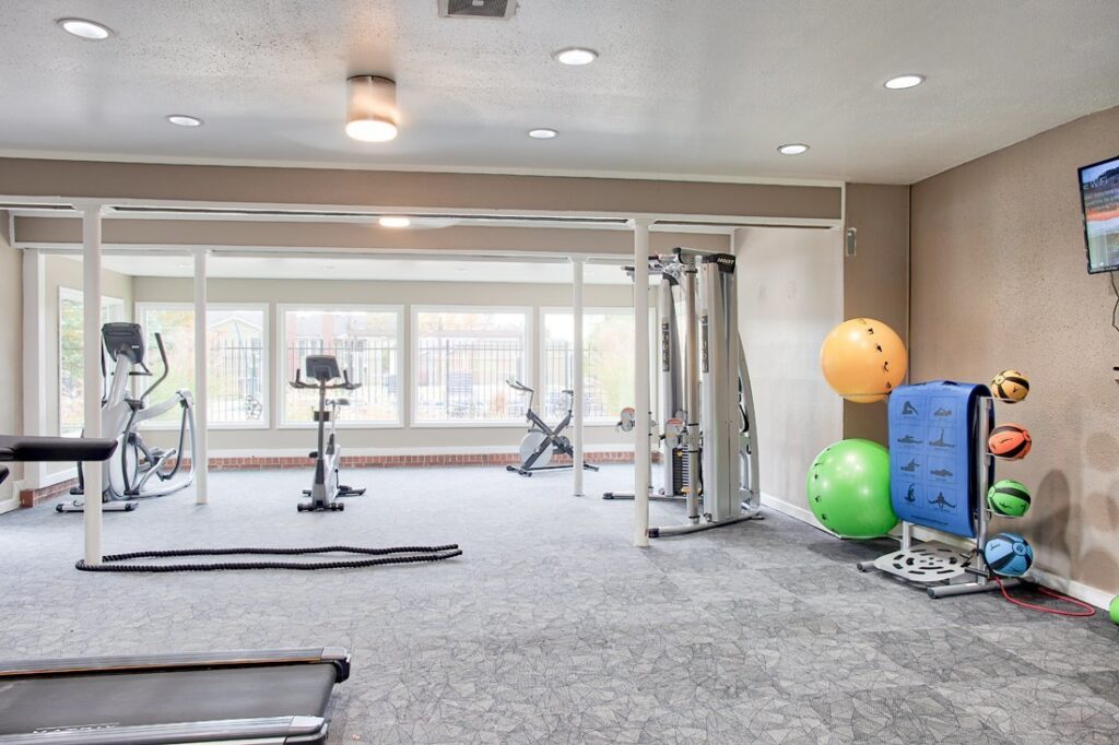 A well-lit gym at Enclave at Eagle Creek features exercise bikes, a treadmill, weight machine, stability balls, and medicine balls, all set near large windows.