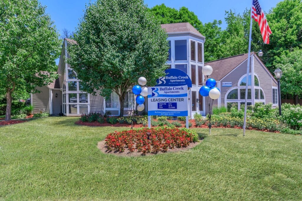 Leasing center for Buffalo Creek Apartments with a sign, balloons, landscaped flower beds, trees, and an American flag in front of a building.