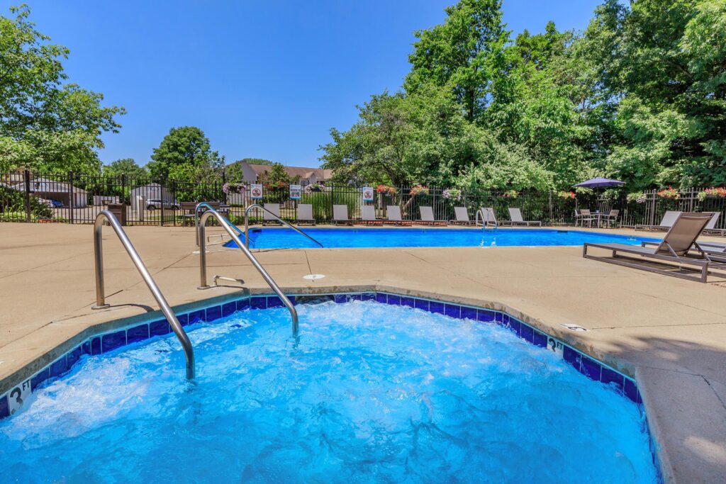 A hot tub with bubbling water in the foreground at Buffalo Creek Apartments, and a swimming pool with lounge chairs and trees in the background on a sunny day.