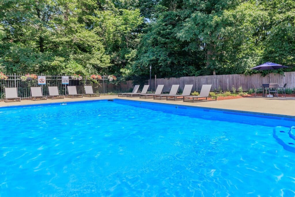 Outdoor swimming pool with clear blue water, surrounded by lounge chairs and trees. A patio table with an umbrella is visible near a wooden fence in the background.