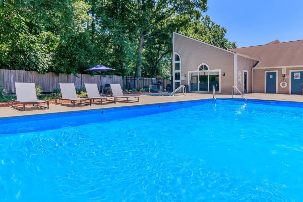 Outdoor swimming pool with bright blue water, several loungers on the deck, shaded patio area, and a building with large windows in the background.