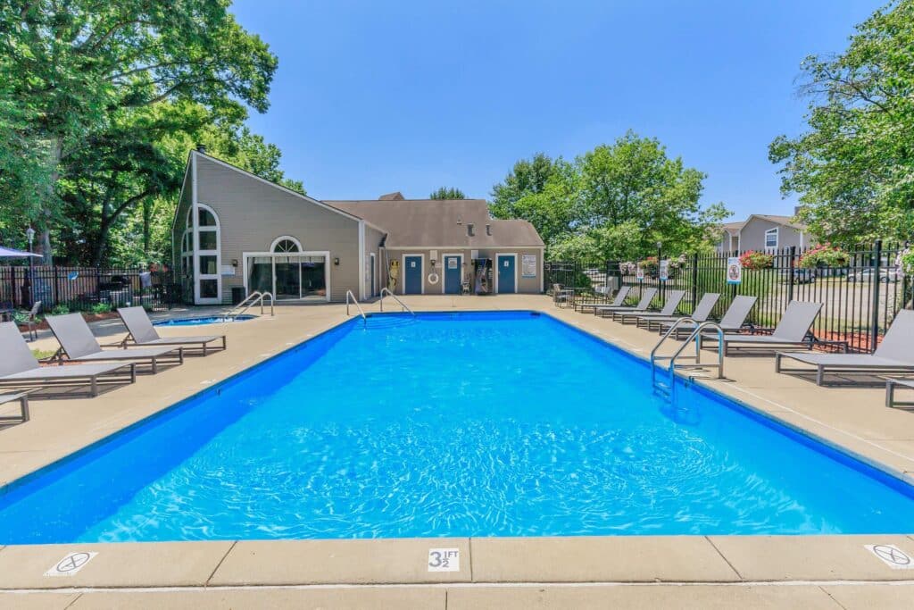 Outdoor swimming pool with clear blue water, surrounded by lounge chairs and trees, with a modern clubhouse building in the background under a clear sky.
