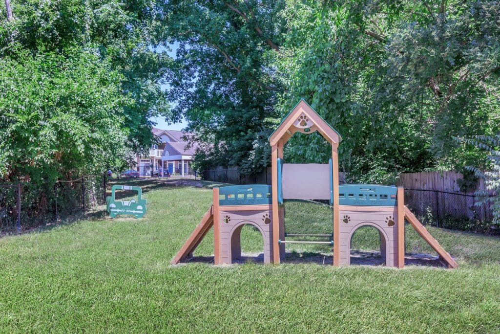 A small playground structure sits on a grassy lawn, surrounded by trees and a fence, with a house visible in the background.