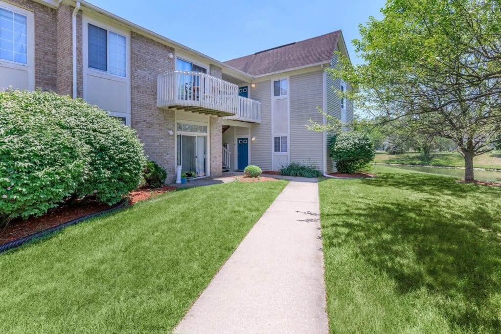 A sidewalk leads to the entrance of a two-story apartment building with brick and siding exterior, surrounded by green lawn and shrubs.