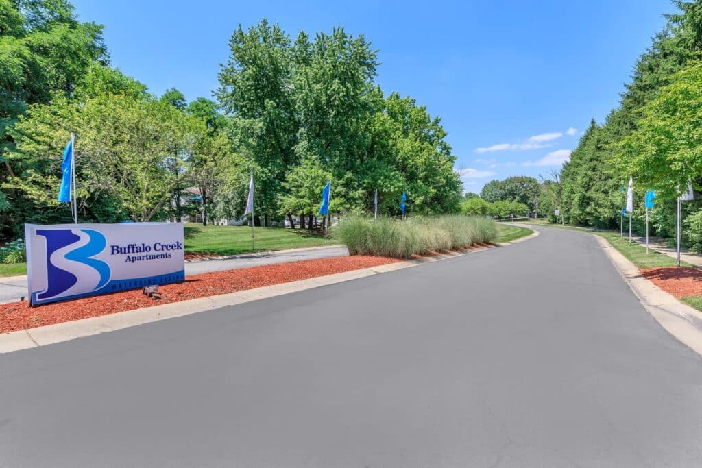Entrance road to Buffalo Creek Apartments with a blue sign on the left, surrounded by trees and landscaping, under a clear sky.