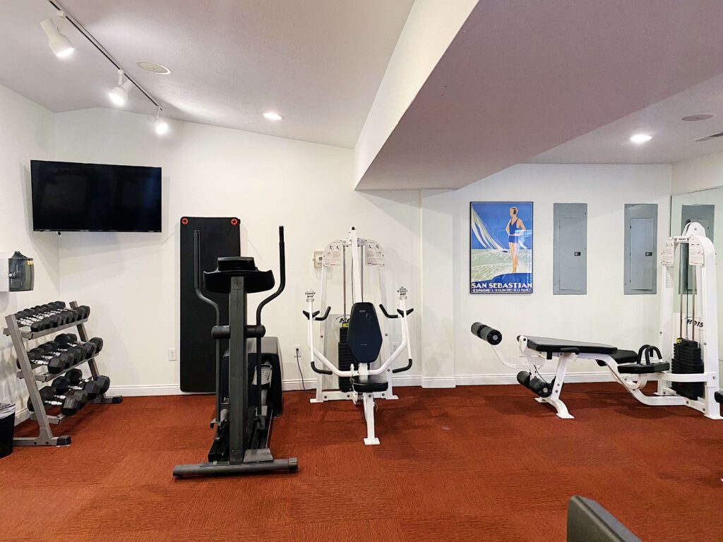 A small gym room with dumbbells, a treadmill, weight machines, a flat-screen TV, and a wall poster on red carpeting.