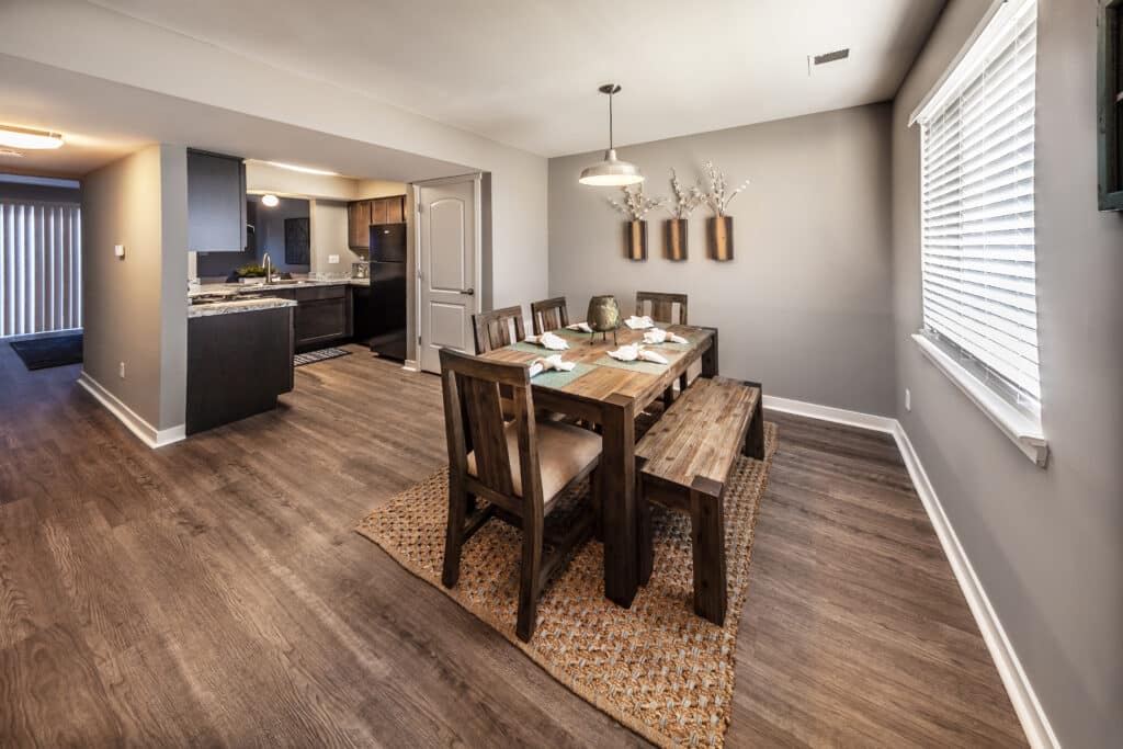 Dining area with wooden table, chairs, and bench on a woven rug at Wyckford Commons Apartments. Kitchen visible in the background. Three wall vases with white branches decorate a gray accent wall.