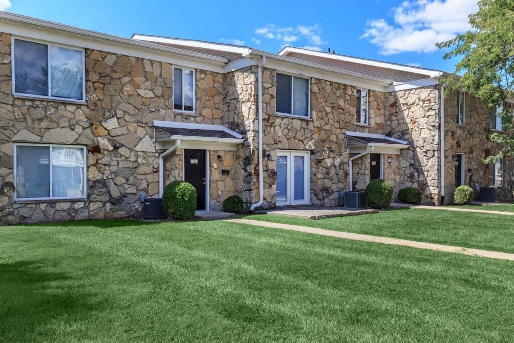 10 West Apartments is a two-story stone building with four units, front-facing windows, shrubs by the entrances, and a well-kept green lawn under a blue sky.