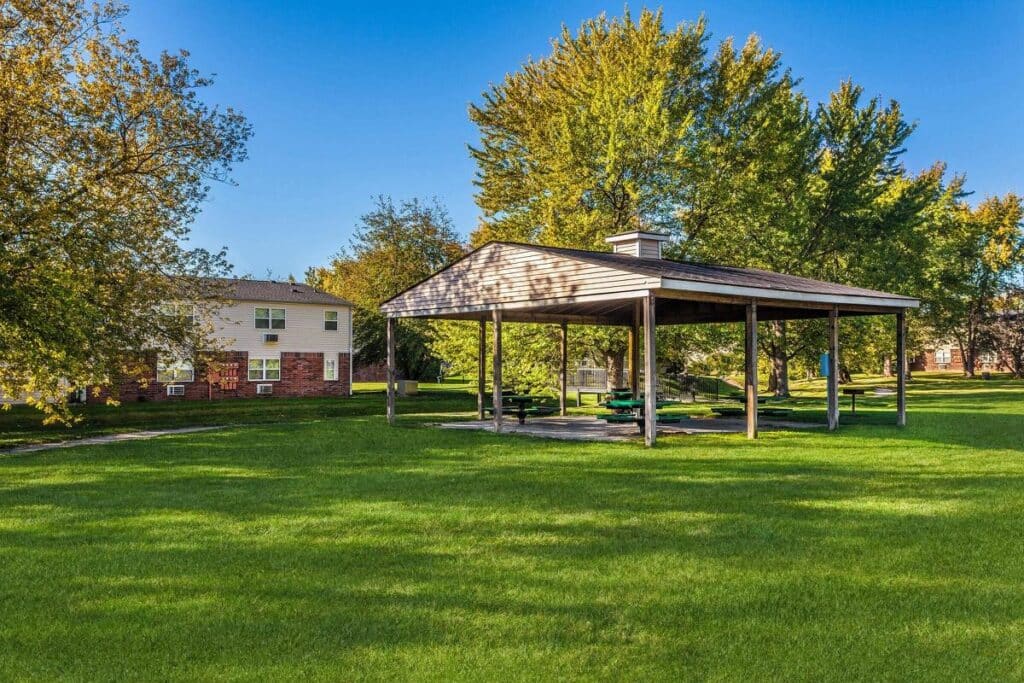 A wooden pavilion with picnic tables stands on a grassy lawn near trees and apartment buildings under a clear blue sky.