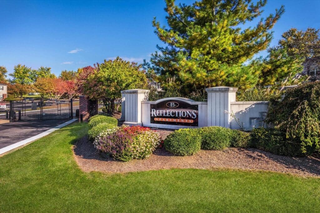 Entrance to Reflections Apartments with a sign, landscaped bushes, and a gated driveway, set against a backdrop of trees and a clear blue sky.