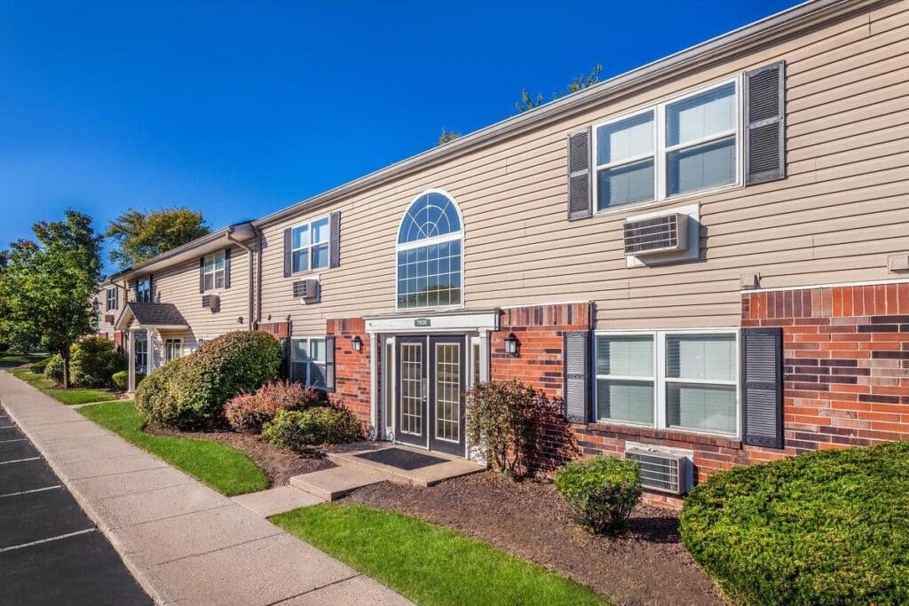 A two-story apartment building with beige siding, brick accents, multiple windows, and landscaped bushes under a clear blue sky.