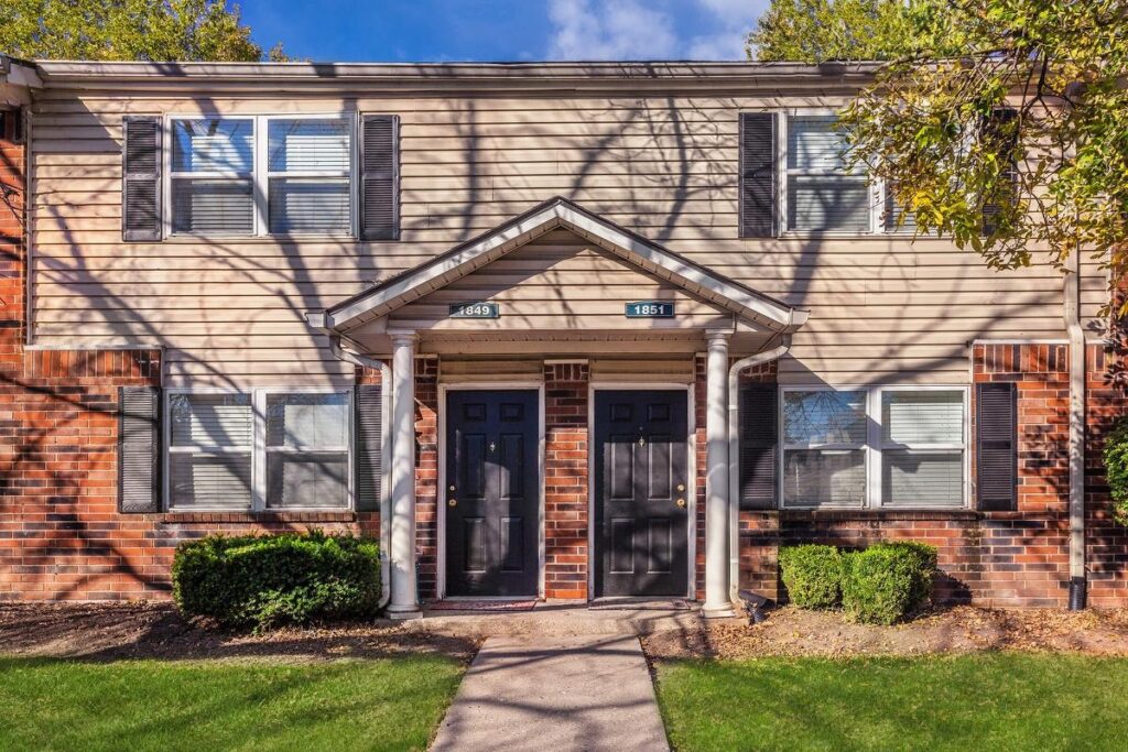 Two-story apartment building with tan siding, brick accents, two black front doors, white columns, and neatly trimmed grass.