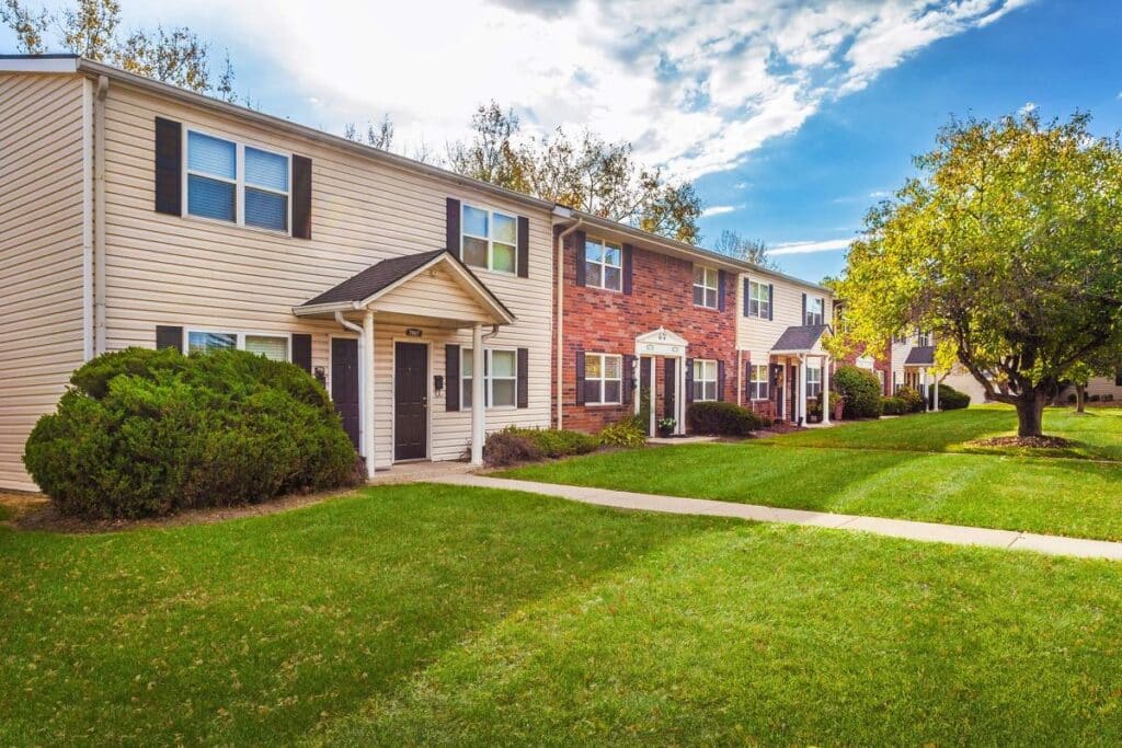 Two-story apartment buildings with siding and brick exteriors are surrounded by green lawns, bushes, trees, and a concrete walkway under a partly cloudy sky.