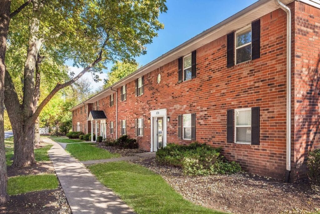 A red brick apartment building with black shutters, surrounded by trees and shrubs, with a sidewalk running alongside it on a sunny day.