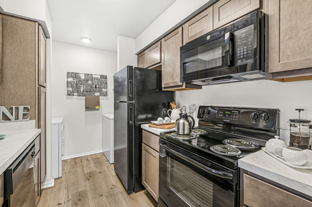 Modern kitchen at Enclave at Eagle Creek features black appliances, wooden cabinets, an electric stove, microwave, refrigerator, and a washer and dryer conveniently visible in the background.