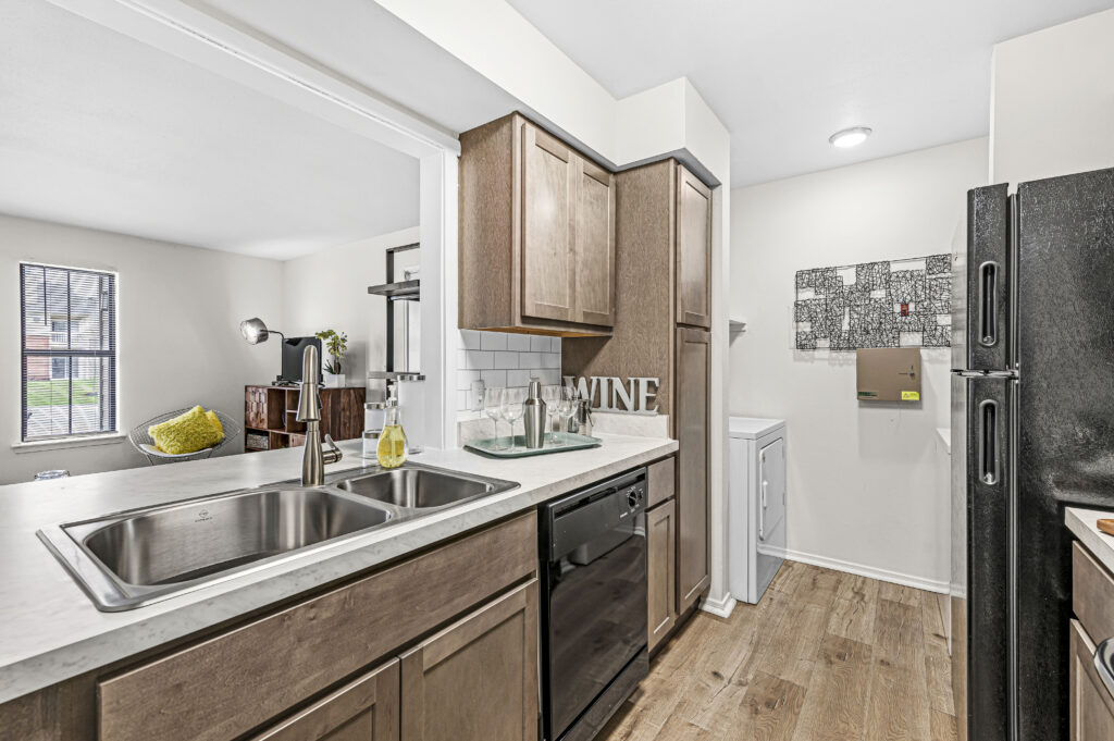 Modern kitchen with wood cabinets, stainless steel sink, black appliances, and a view into the living room at Enclave at Eagle Creek; laundry machines visible in a small adjacent area.