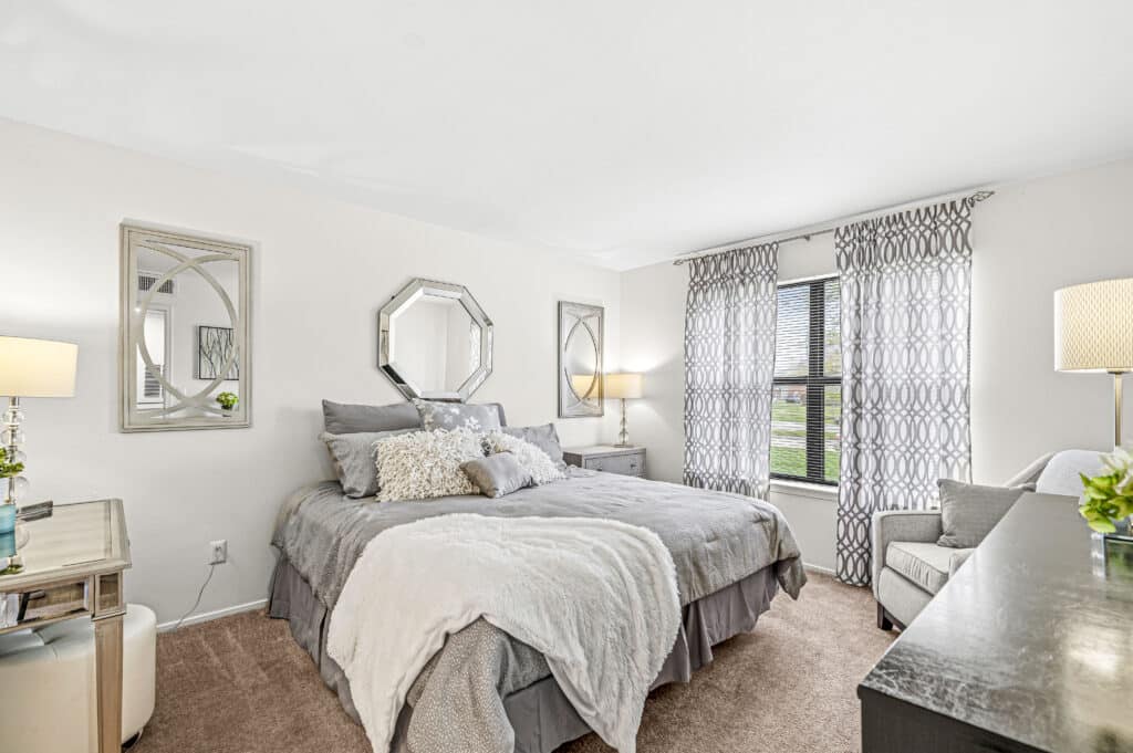 A bedroom at Enclave at Eagle Creek features a neatly made bed, two bedside tables with lamps, a desk with a chair, and two large windows adorned with patterned curtains.